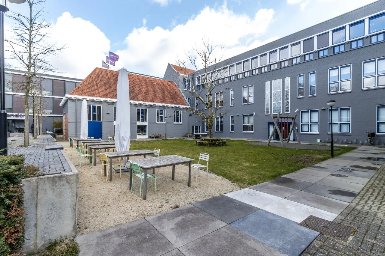 Binnenplein van een modern grijs gebouw aan de Lage Naarderweg met picknicktafels, kleurrijke stoelen en een boom in het gras.